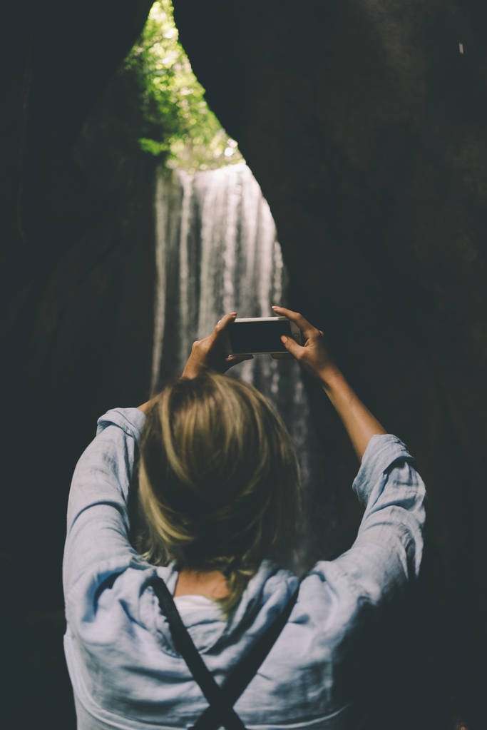 Widok z tyłu zdjęcie Kobieta blonde biorąc zdjęcie wybitne piękny wodospad przez aparat jej smartphone w głąb tropikalnej dżungli podczas letnich wakacji.  - Zdjęcie, obraz