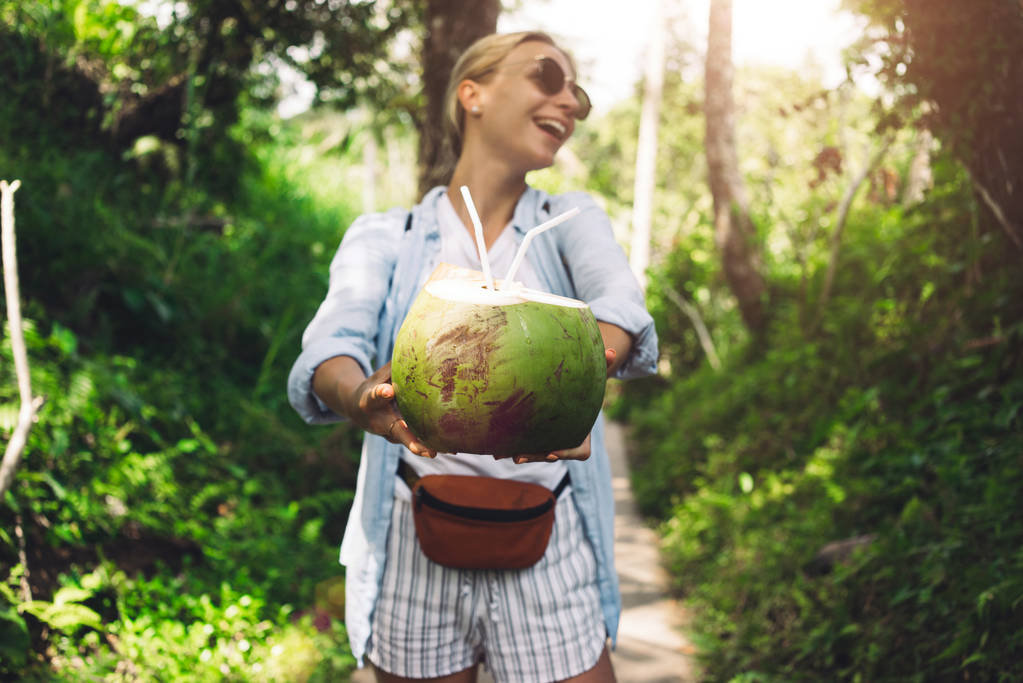 Dziewczyna Blonde turystycznych oferuje kokos koktajl na tle zielonej dżungli. Zielony kokos z słomki w ręce kobiety roześmiany spacer w lesie tropikalnym podczas letnich wakacji. - Zdjęcie, obraz