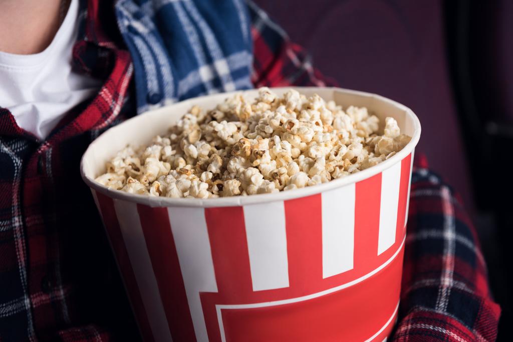 обрізаний вид людини, що тримає великий кошик попкорну в кінотеатрі
 - Фото, зображення