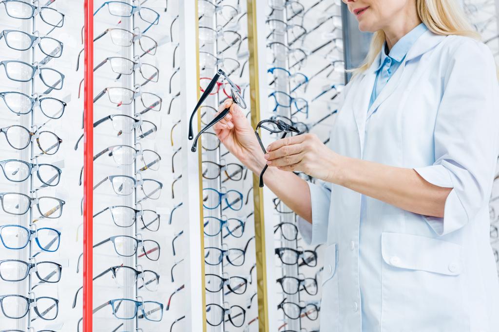 Ausgeschnittene Ansicht einer professionellen Augenärztin, die in Optik mit Brille im Regal steht - Foto, Bild