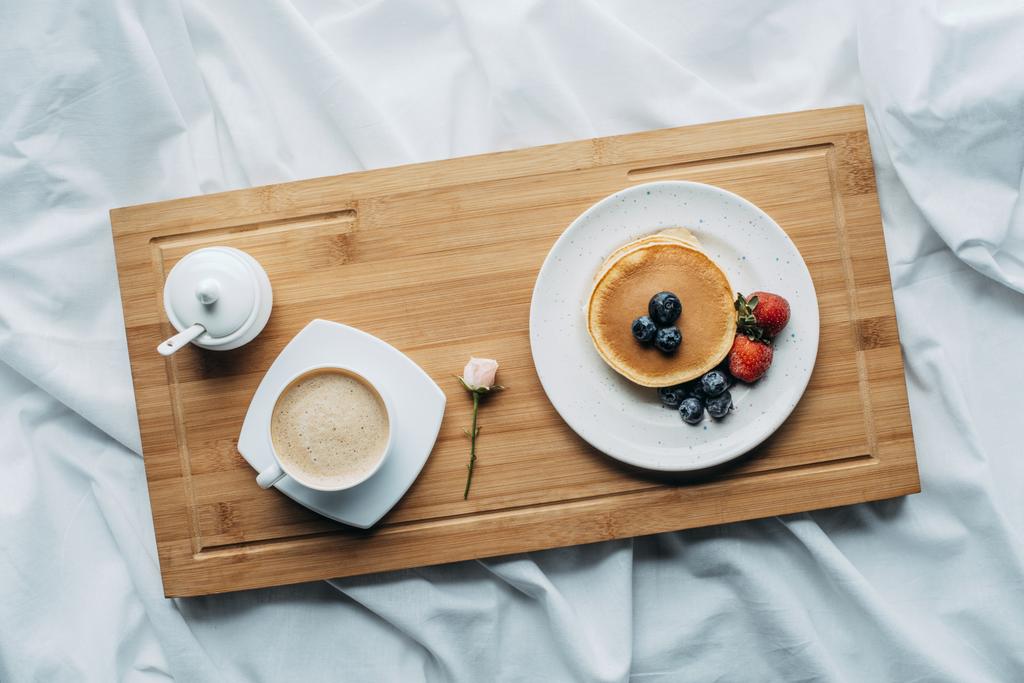 bovenaanzicht van een ontbijt op bed met lekkere pannenkoeken en koffie op houten dienblad - Foto, afbeelding