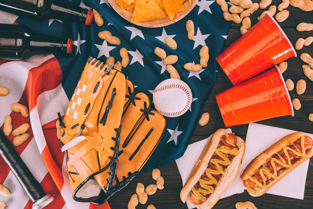 вид зверху на хот-доги, пластикові чашки, арахіс, пляшки пива, бейсбольний м'яч і рукавички з кажаном на американському прапорі
 - Фото, зображення