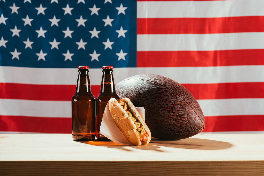 крупним планом пляшки пива, хот-дог і м'яч регбі на дерев'яному столі з нами прапор позаду
 - Фото, зображення