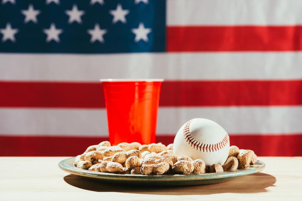 Nahaufnahme von Baseballball auf Teller mit Erdnüssen, roter Plastikflasche und amerikanischer Flagge dahinter - Foto, Bild
