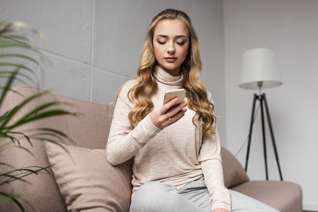 beautiful young woman using smartphone while witting on cozy couch at home - Photo, Image