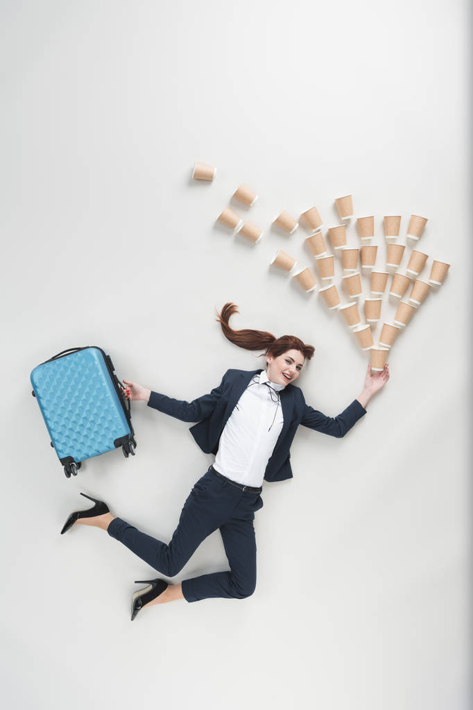 Über-Kopf-Ansicht einer fröhlichen Geschäftsfrau mit Koffer und Einwegbechern isoliert auf grau - Foto, Bild