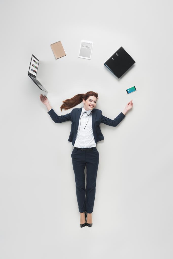 над головой вид бизнесвумен с различными офисными принадлежностями с помощью ноутбука, изолированного на сером
 - Фото, изображение