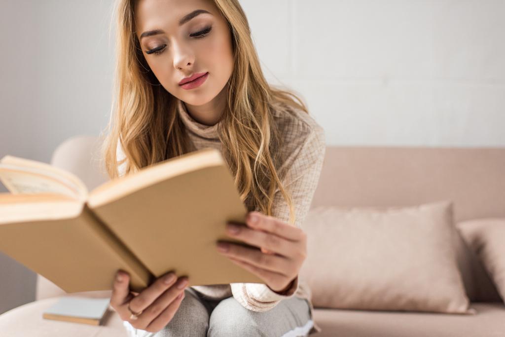 attractive young woman reading book on cozy couch at home - Photo, Image