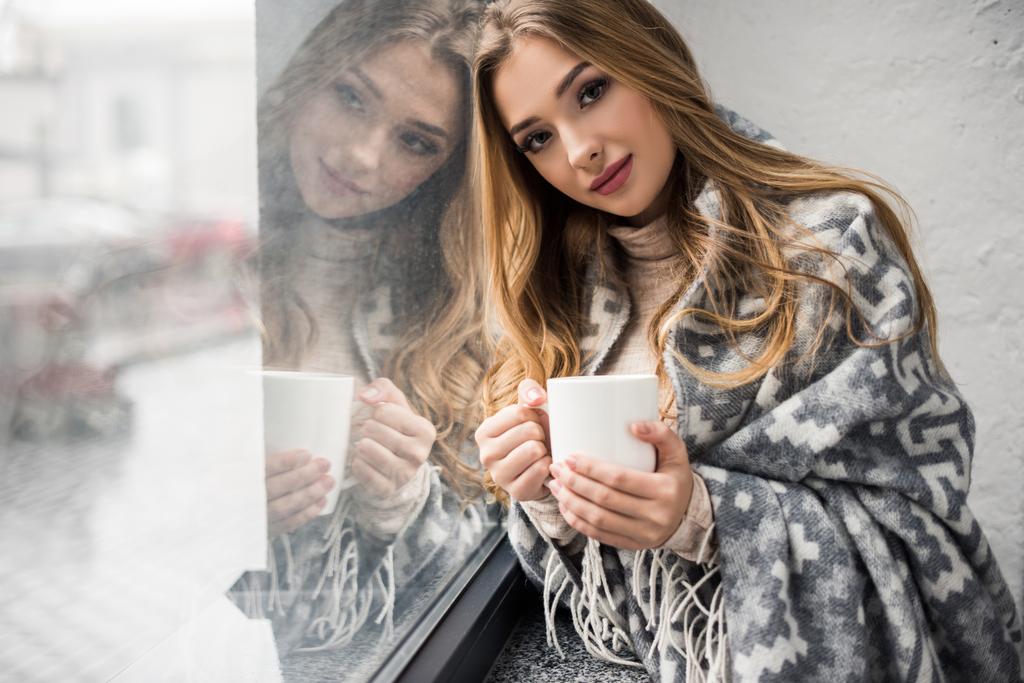 mooie jonge vrouw drinken koffie terwijl leunend op venster - Foto, afbeelding