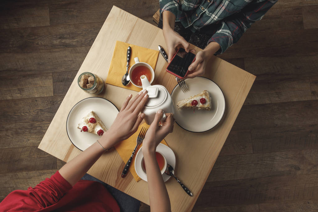 vista superior de la persona que vierte el té de la tetera mientras que amigo usando el teléfono inteligente durante el desayuno
 - Foto, Imagen