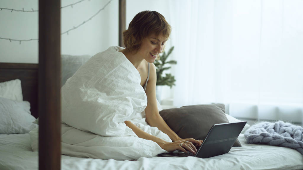 Veselá mladá žena pomocí její přenosný počítač seděl v posteli doma - Fotografie, Obrázek