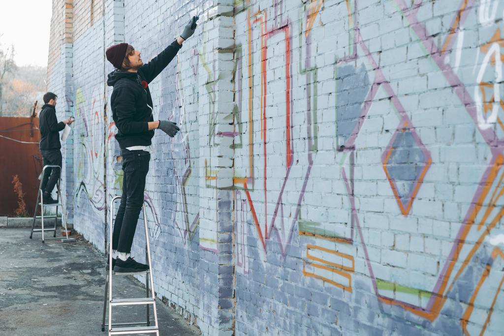 katutaiteilijat seisovat tikkailla ja maalaavat värikkäitä graffiteja rakennukseen
 - Valokuva, kuva