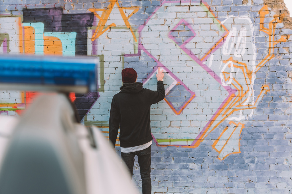 Rückansicht von Vandalen, die Graffiti an Wand malen, Polizeiauto im Vordergrund - Foto, Bild