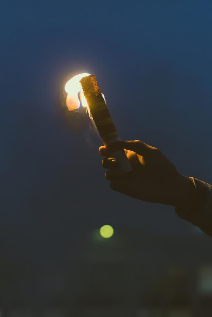 обрезанный вид мужской руки с дымовой бомбой с огнем ночью
 - Фото, изображение