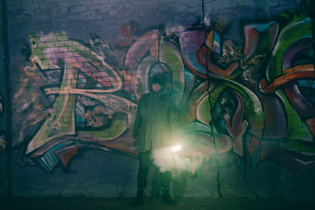 Mann hält Rauchbombe in der Hand und steht nachts mit Graffiti gegen Wand - Foto, Bild