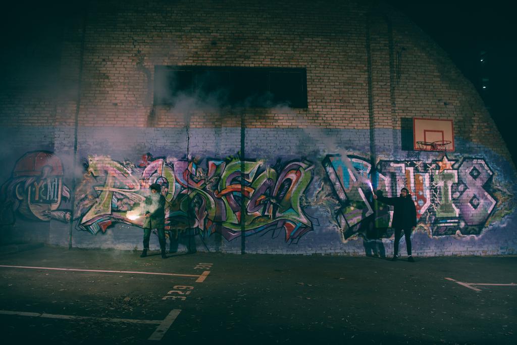 people holding smoke bombs and standing against wall with graffiti at night - Photo, Image