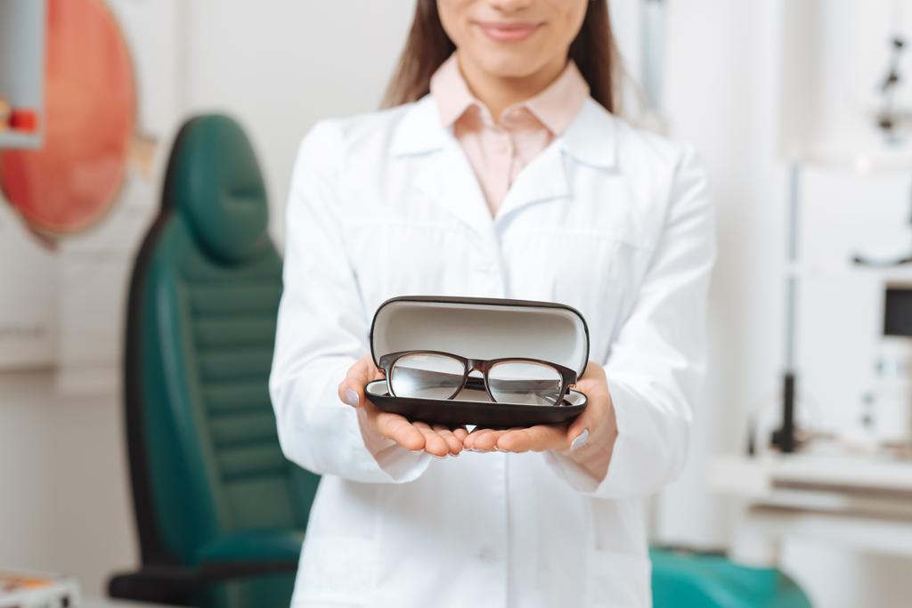 partial view of optician in white coat holding pair of eyeglasses in clinic - Photo, Image
