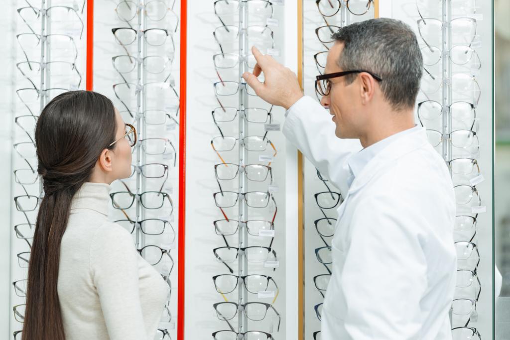 vista lateral del oculista ayudando a la mujer a elegir un par de gafas en óptica
 - Foto, imagen