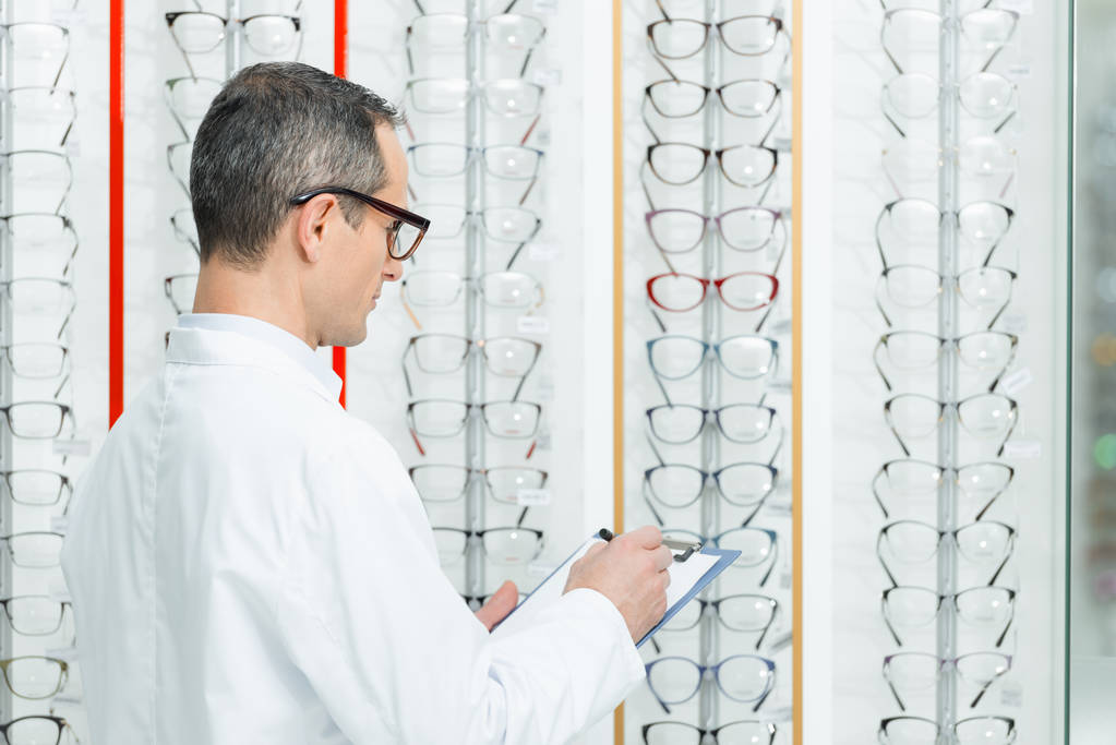 vue latérale de l'optométriste prenant des notes tout en se tenant sur des étagères avec des lunettes en optique
 - Photo, image