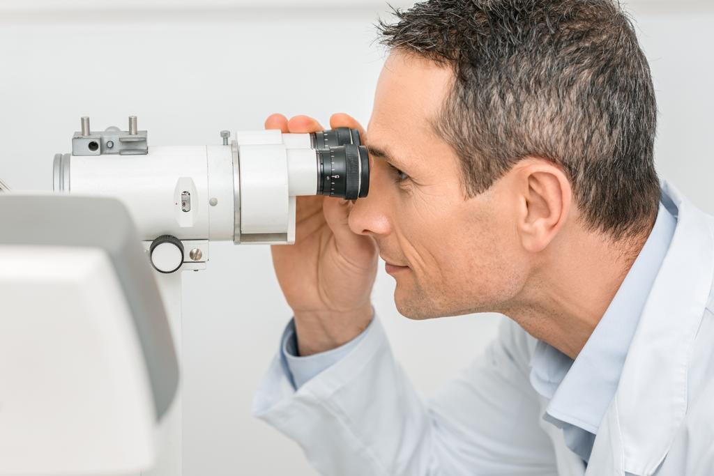 vista lateral del optometrista profesional que trabaja con lámpara de hendidura en clínica
 - Foto, Imagen