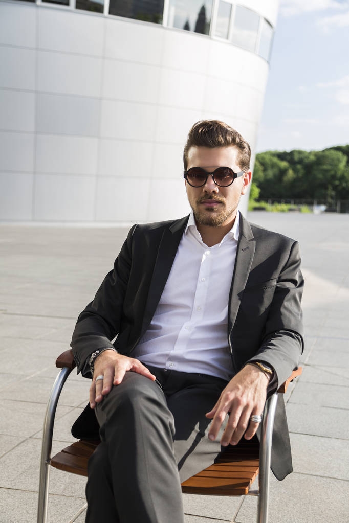 Mannelijke zakenman of werknemer in zwart pak zitten op stoel in de buurt van modern gebouw - Foto, afbeelding