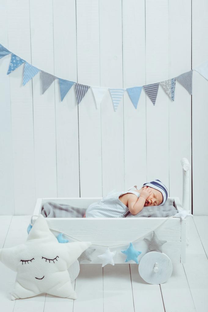 schattige pasgeboren baby in hoed slapen in houten kinderbed met ster kussen in de buurt van door  - Foto, afbeelding