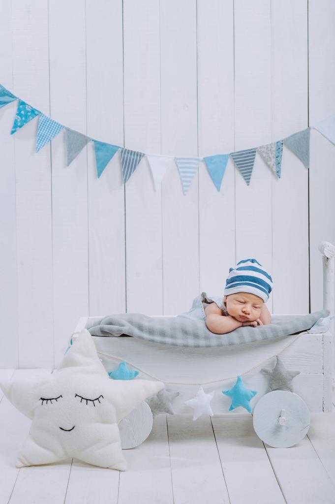 милий новонароджений дитина в капелюсі спить в дерев'яному дитячому ліжечку з зірковою подушкою поруч
  - Фото, зображення