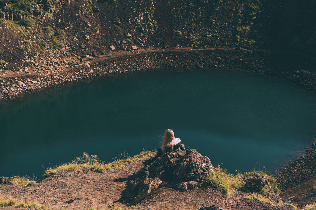 Rückansicht einer jungen Frau, die auf einem Felsen sitzt und auf einen schönen Kratersee in Island blickt  - Foto, Bild