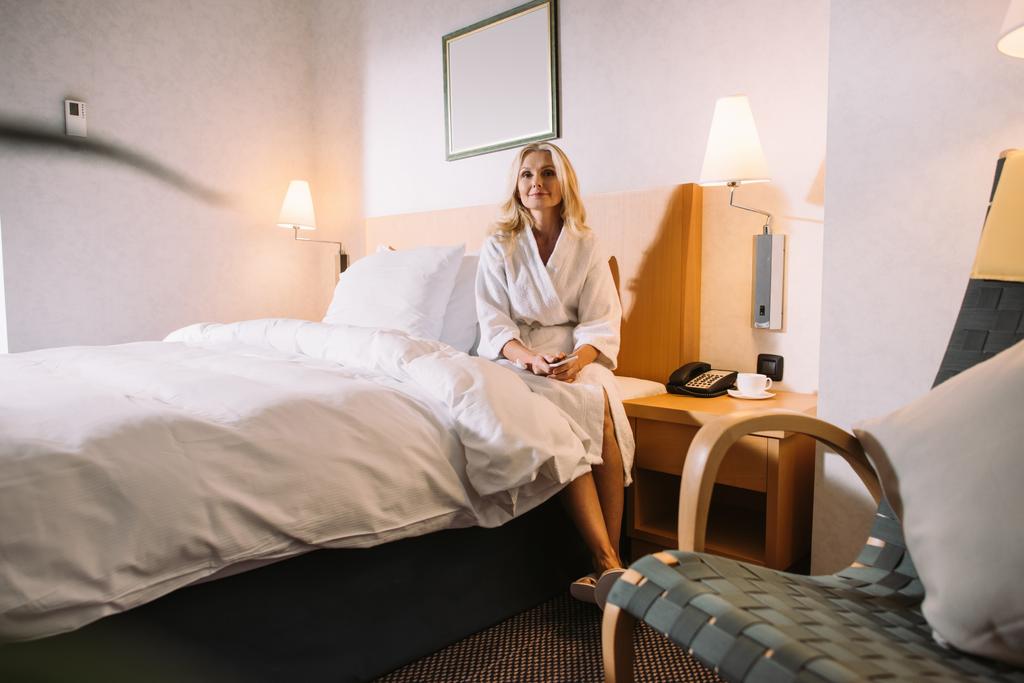 rijpe vrouw met smartphone in handen zittend op bed en op zoek weg in hotelkamer - Foto, afbeelding