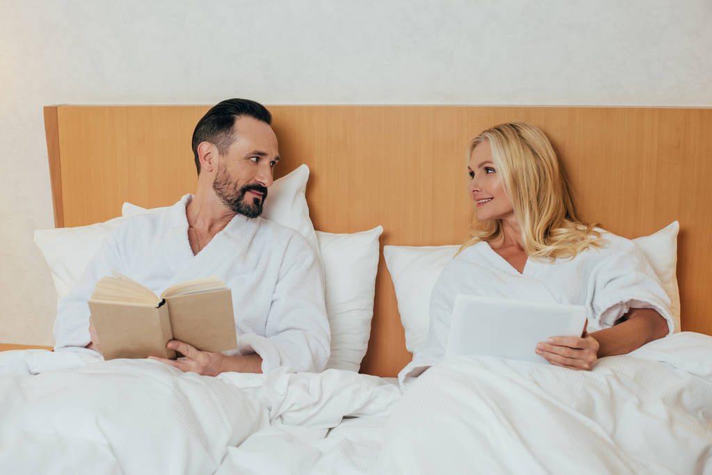 зріла пара з цифровим планшетом і книгою посміхається один одному, лежачи в ліжку в готельному номері
 - Фото, зображення