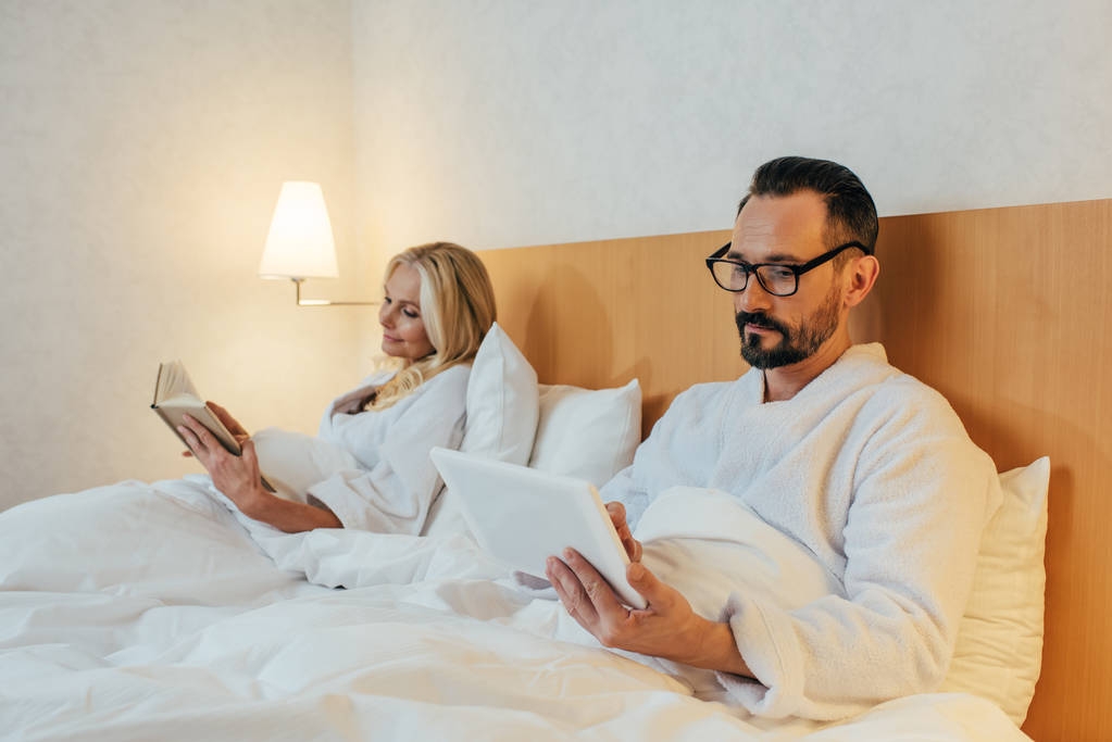 połowie para dorosłych w szlafroki, czytając książkę i za pomocą cyfrowego tabletu leżąc w łóżku w pokoju hotelowym - Zdjęcie, obraz