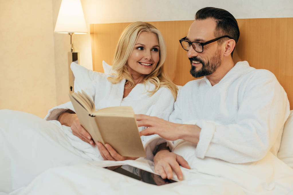 Lächelndes Paar mittleren Alters liest Buch und nutzt digitales Tablet, während es zusammen im Hotelzimmer im Bett liegt - Foto, Bild