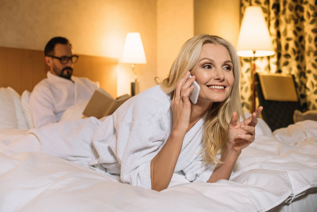 mooie rijpe vrouw in badjas liggend op bed en praten over smartphone terwijl echtgenoot leesboek achter  - Foto, afbeelding