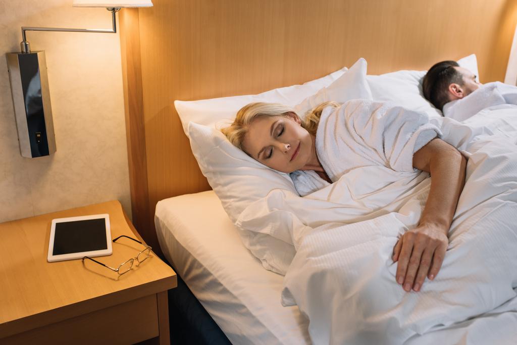 dospělý pár v župany spí v posteli a digitální tablet s brýlemi na stole v hotelovém pokoji  - Fotografie, Obrázek