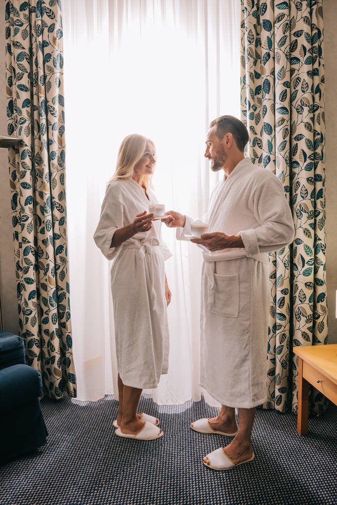 полный вид счастливой зрелой пары в халатах, пьющей кофе и улыбающейся друг другу в гостиничном номере
   - Фото, изображение