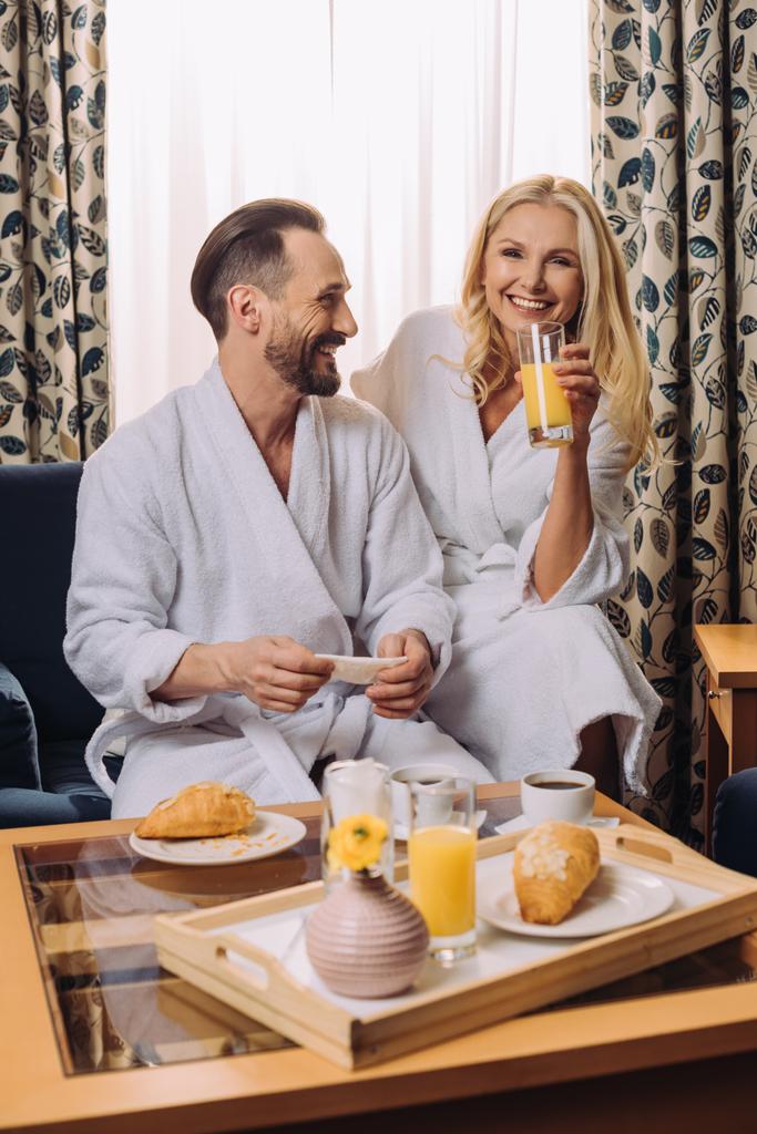 ευτυχισμένο μέσης ηλικίας ζευγάρι σε μπουρνούζια έχοντας πρωινό μαζί σε δωμάτιο ξενοδοχείου  - Φωτογραφία, εικόνα
