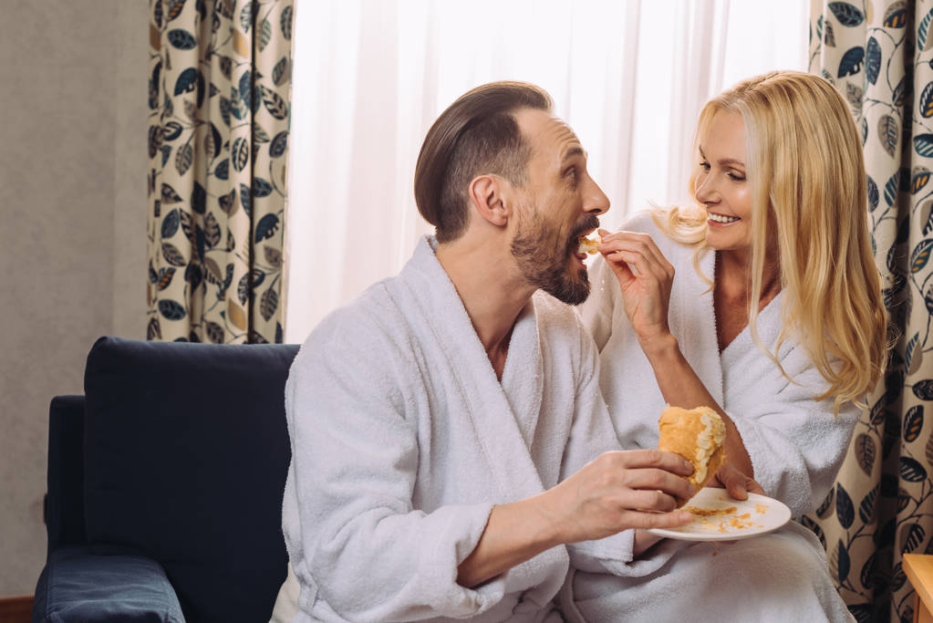feliz mujer madura en albornoz alimentar a su marido con pastelería durante el desayuno en la habitación de hotel
  - Foto, imagen