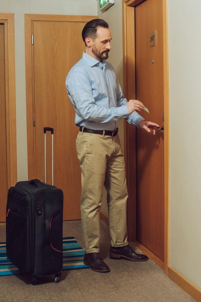 bärtiger Mann mit Koffer mit Karte und sich öffnender Hoteltür - Foto, Bild