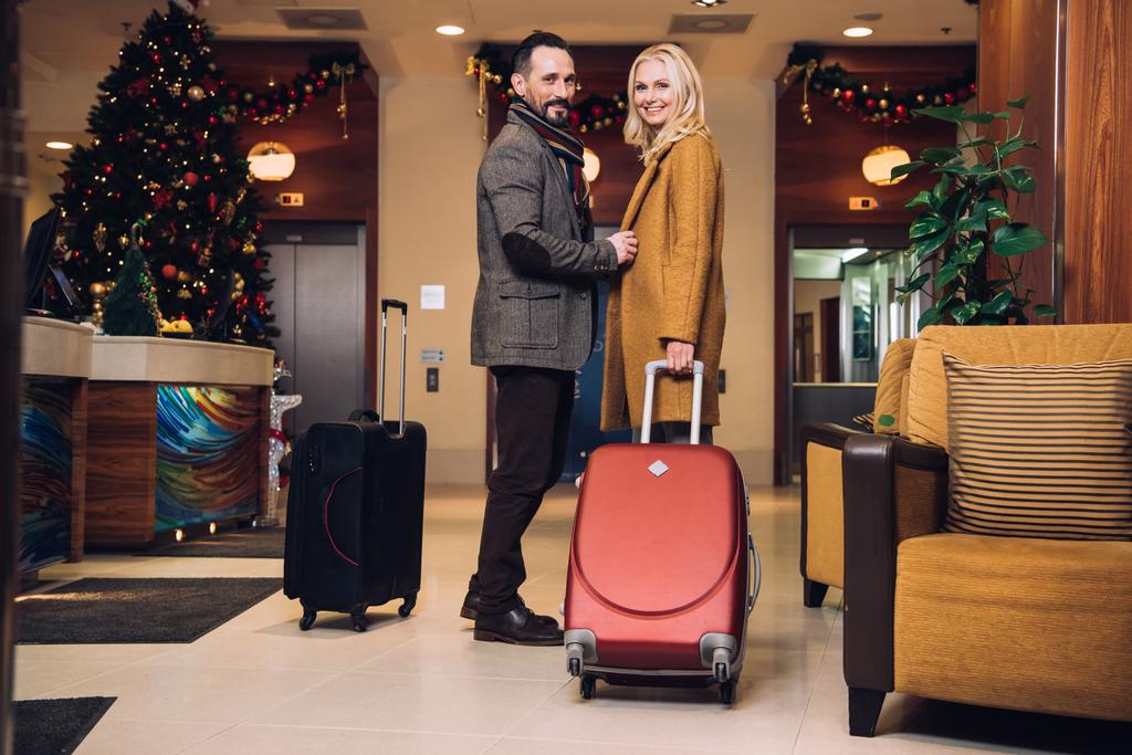 スーツケースと中年夫婦の地位とクリスマスにホテルの廊下でカメラに笑顔 - 写真・画像