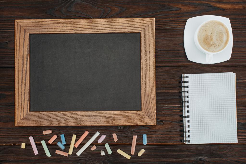 flat lay com quadro branco vazio, xícara de café, caderno em branco e giz em mesa de madeira escura
 - Foto, Imagem