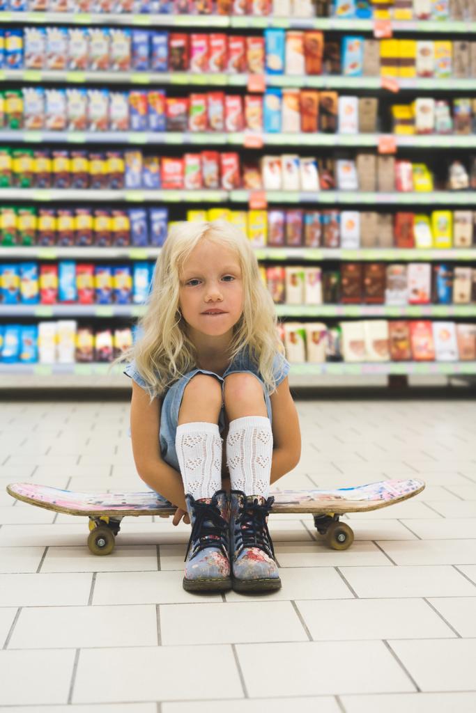 rozkošná blondýnka dítě sedí na skateboardu v supermarketu s police za - Fotografie, Obrázek