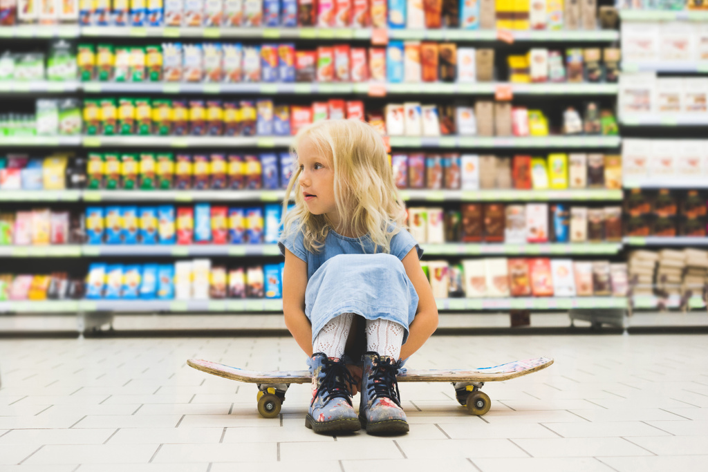 little blonde child sitting on skateboard in supermarket with shelves behind - Photo, Image