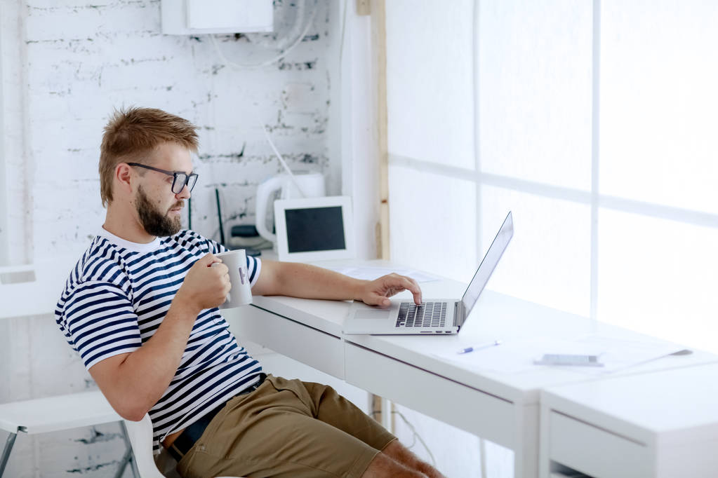 joven hombre de negocios guapo que trabaja en el ordenador portátil sentado en el escritorio y beber café en la oficina
 - Foto, imagen