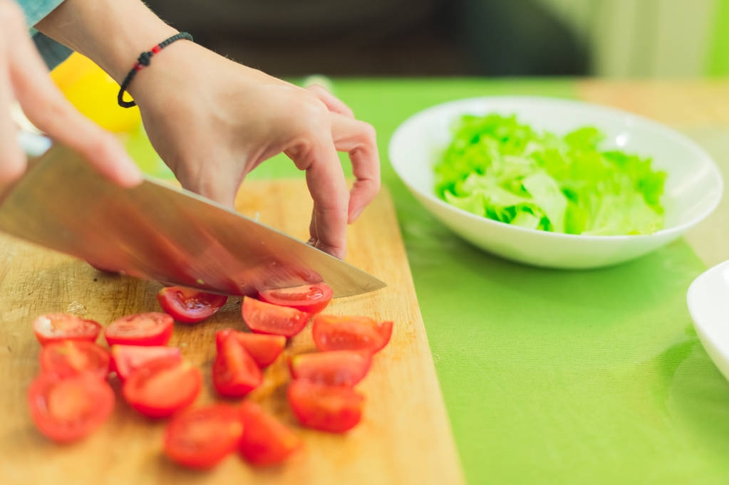 Las manos de una joven cortan los tomates cherry en una tabla de cortar de madera sobre una mesa verde en un entorno hogareño
 - Foto, Imagen