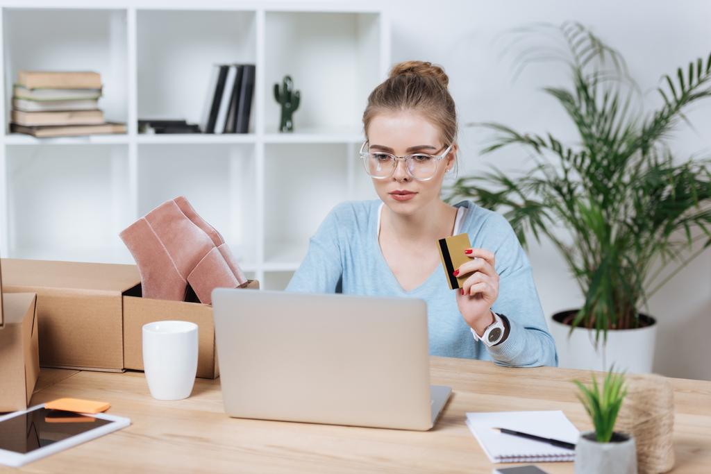 портрет сфокусированной женщины с кредитной картой в руке, смотрящей на экран ноутбука за столом
 - Фото, изображение