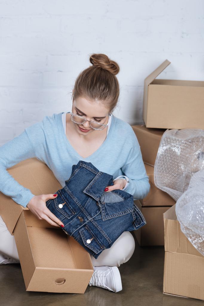 mladý podnikatel uvedení džínové bundy do lepenkové krabice, zatímco balení výrobků na domácí kancelář - Fotografie, Obrázek