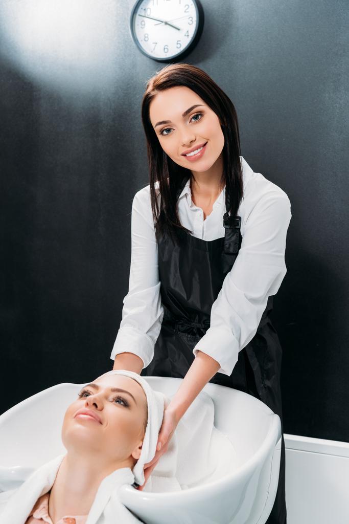 smiling hairdresser drying washed customer hair with towel and looking at camera - Photo, Image