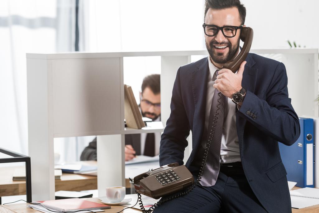 бізнесмен розмовляє стаціонарним телефоном в офісі і сидить на столі
 - Фото, зображення