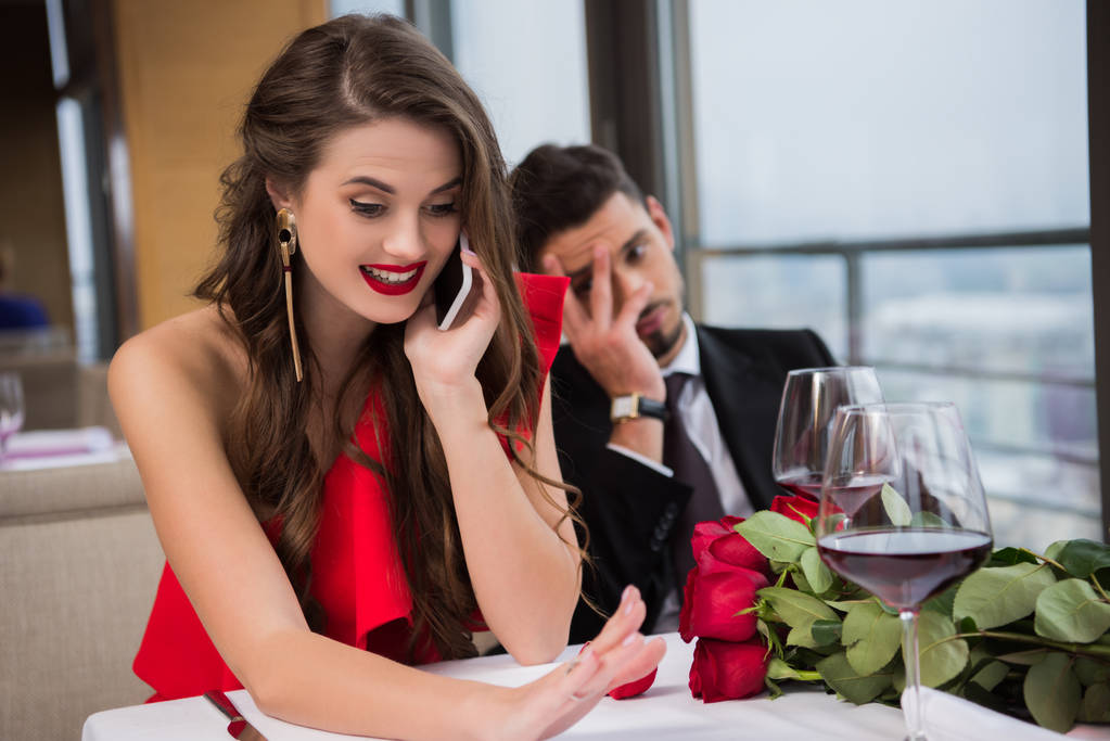 mutlu nişanlısı Smartphone'da yakınındaki restoranda oturan bıkkın erkek arkadaşı ile konuşurken seçici odak - Fotoğraf, Görsel
