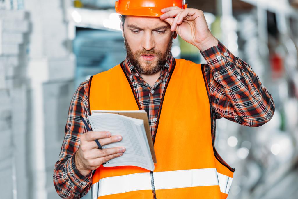 pensive worker in safety vest looking on notepad in storehouse - Photo, Image